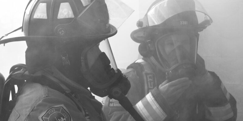 Two female firefighters in full turnout gear adjust their breathing apparatus in a smoky room.